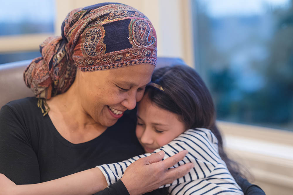 A beautiful senior woman with cancer holds her granddaughter tightly in her lap. Grandma is wearing a headscarf and smiling.
