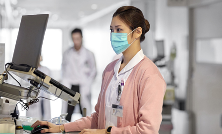 Female nurse using laptop while working on medical reports. Healthcare professional is busy in hospital. She is wearing surgical mask and uniform.