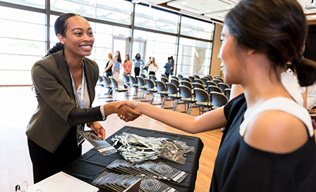 Cheerful mid adult businesswoman shakes hands with a colleague during a conference. The businesswoman is working at the conference registration table.