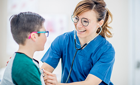 A woman holds a stethoscope to a young caucasian boy's heart, while they exchange smiles. They are both wearing eyeglasses and they are sitting in a doctor's office.