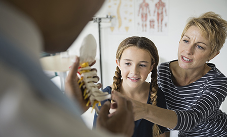 Mother and daughter discussing the human spine with a doctor, who holding a model of the human spine.
