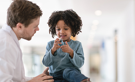 A black toddler boy sits on an examination table while at a medical consultation. The child has asthma and is learning to use an inhaler. The boy is holding a puffer and the doctor is explaining how to use the device.